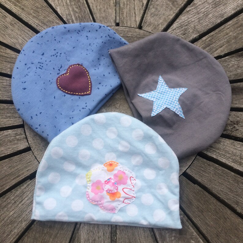Op de afbeelding: Drie baby mutsen met verschillende patronen en applicaties. E&eacute;n is blauw met witte stippen en een bruin hart, &eacute;&eacute;n is grijs met een blauw-witte geruite ster en &eacute;&eacute;n is lichtblauw met witte stippen en een roze-oranje bloem.