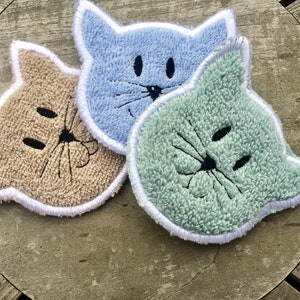 May include: Three cat-shaped kitchen scrubbers in blue, beige, and green. Each scrubber has a white border and embroidered black eyes, nose, and whiskers.