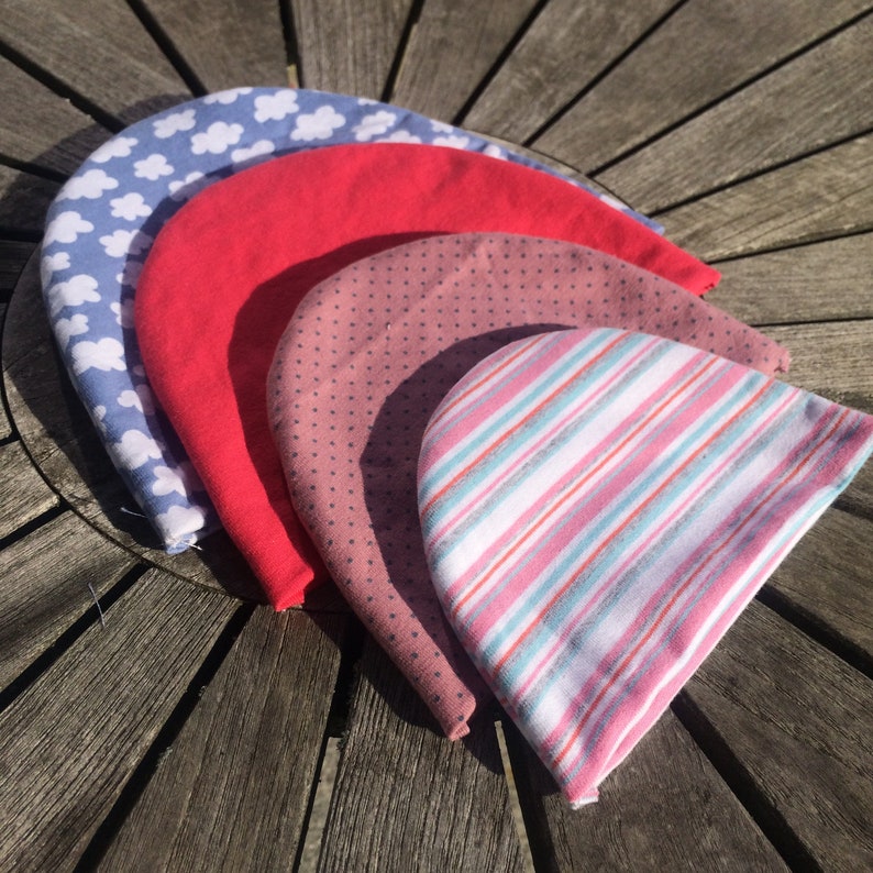 May include: Four knitted beanies in different colours and patterns. The beanies are stacked on top of each other, with the blue beanie with white clouds on top, followed by a red beanie, a pink beanie with small white dots, and a white beanie with pink and blue stripes.