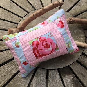 Könnte beinhalten: Ein rosa-blaues Patchwork-Kissen mit einer großen rosa Rose in der Mitte. Das Kissen ist mit weißer Spitzenborte verziert und hat ein gestepptes Design.