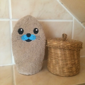 May include: A brown washcloth with an embroidered seal face. The seal has black eyes, a blue nose, and black whiskers. The washcloth is sitting next to a woven wicker basket with a lid.