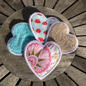 May include: Three heart-shaped fabric pads, one blue, one beige, and one pink with a floral pattern. The pads are on a wooden surface.