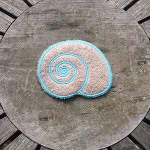 May include: A beige and blue embroidered seashell with a spiral design. The seashell is on a wooden surface.