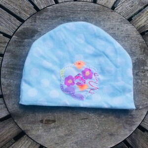 Op de afbeelding: Een lichtblauwe baby muts met witte stippen en een kleurrijke bloemenapplicatie. De bloem heeft een roze, paarse, oranje en gele ontwerp met een kleine knop in het midden.