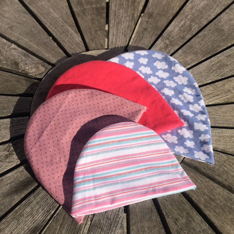 May include: Four different fabric beanies stacked on top of each other. The top beanie is blue with white cloud designs. The second beanie is red. The third beanie is pink with brown polka dots. The bottom beanie is white with pink and blue stripes.