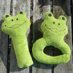 May include: Two plush green frog toys. One frog is a long, thin toy with a smiling face. The other frog is a ring toy with a smiling face.