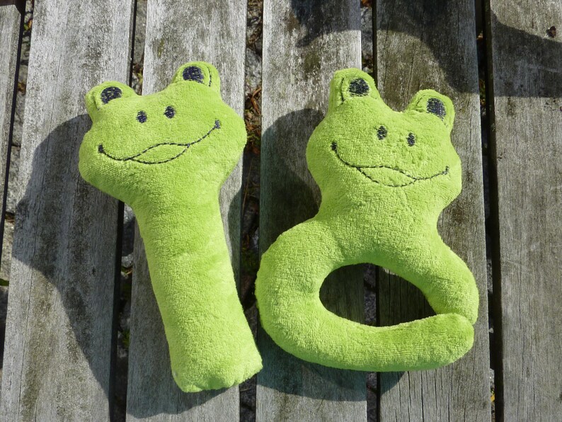May include: Two plush green frog toys. One frog is a long, thin toy with a smiling face. The other frog is a ring toy with a smiling face.