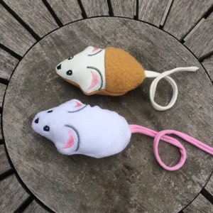 May include: Two plush cat toys shaped like mice. One mouse is white with brown fur on the back and a white tail. The other mouse is white with a pink tail.