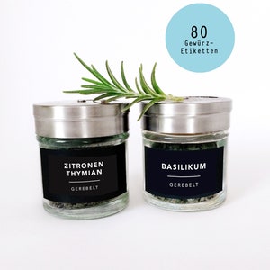 May include: Two glass spice jars with silver lids and black labels. One jar is labelled "Zitronen Thymian" and the other "Basilikum". A sprig of rosemary rests on top. A light blue circle with the text "80 Gewürz-Etiketten" is in the background.