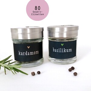 Könnte beinhalten: Zwei gläserne Gewürzgläser mit Metalldeckeln und schwarzen Etiketten. Die Etiketten haben weißen Text und ein kleines Herzsymbol. Die Gläser sind mit getrockneten Kräutern gefüllt. Die Etiketten lauten "Kardamom" und "Basilikum".