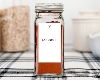 May include: A clear glass spice jar with a silver lid, filled with red-orange tandoori spice. A white label with a red heart and the word "TANDOORI" is affixed to the front. The jar sits on a black and white checkered surface.