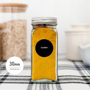 Könnte beinhalten: Ein klares Glasgewürzglas gefüllt mit gelbem Currypulver. Das Glas hat einen silbernen Deckel und ein schwarzes rundes Etikett mit der Aufschrift "CURRY" in Weiß. Das Etikett hat einen Durchmesser von 30 mm. Das Glas steht auf einer schwarz-weiß karierten Oberfläche.
