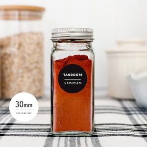 Könnte beinhalten: Ein klares Gewürzglas mit silbernem Deckel, gefüllt mit rotem Tandoori-Gewürz. Ein schwarzes Etikett mit weißem Text lautet "TANDOORI" und "GEMAHEN". Das Glas hat einen Durchmesser von 3 cm. Das Glas steht auf einer schwarz-weiß karierten Oberfläche.