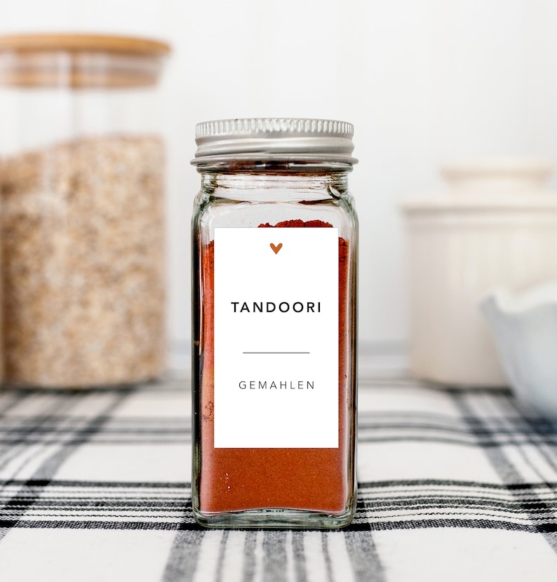 May include: A glass spice jar with a silver lid, filled with red-orange tandoori spice. A white label displays the word "TANDOORI" and "GEMAHEN" in black text, with a small heart symbol. The jar sits on a black and white checkered surface.