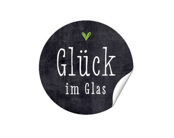 May include: A round, black sticker with the German text "Glück im Glas" in white script, meaning "Happiness in a Glass." A small green heart is above the word "Glück." The sticker has a distressed texture and a corner is peeled back.