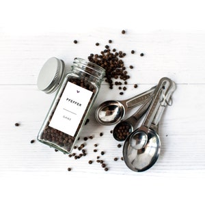 May include: A clear glass jar with a silver lid, filled with black peppercorns, and a white label that reads "PFEFFER GANZ". Scattered peppercorns and a set of stainless steel measuring spoons are also visible.