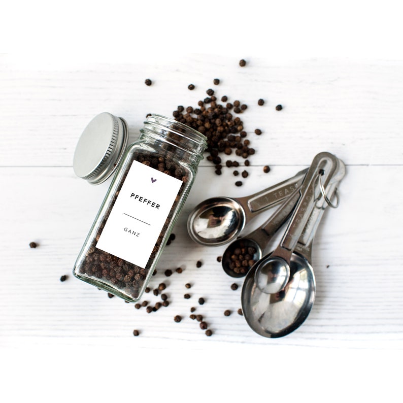 May include: A clear glass jar with a silver lid, filled with black peppercorns, and a white label that reads "PFEFFER GANZ". Scattered peppercorns and a set of stainless steel measuring spoons are also visible.