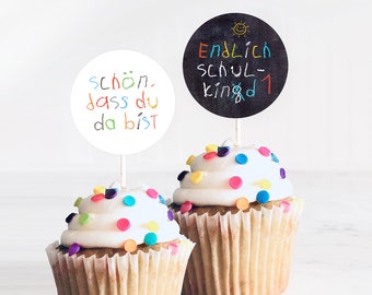 May include: Two cupcakes with white frosting and colorful sprinkles. One cupcake has a round, white sticker with the text "schon, dass du da bist" in colorful handwriting. The other cupcake has a round, black sticker with the text "Endlich schul- kind 1" in colorful handwriting.