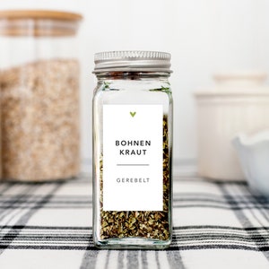 May include: A clear glass spice jar with a silver lid, filled with dried herbs. A white label features the text "Bohnen Kraut" and "Gerebelt" in black, with a green heart above. The jar sits on a black and white checkered surface.