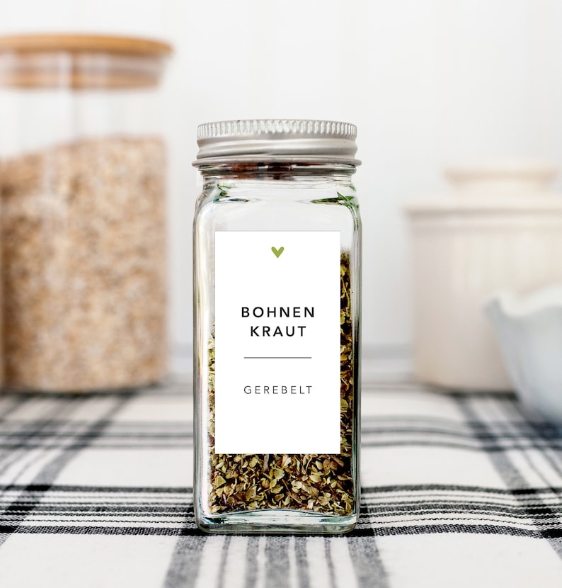 May include: A clear glass spice jar with a silver lid, filled with dried herbs. A white label features the text "Bohnen Kraut" and "Gerebelt" in black, with a green heart above. The jar sits on a black and white checkered surface.