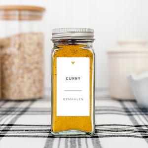 May include: A clear glass spice jar filled with yellow curry powder. The jar has a silver lid and a white label with the word "CURRY" and "GEMAHEN" printed in black. A small gold heart is above the word "CURRY". The jar sits on a black and white checkered surface.
