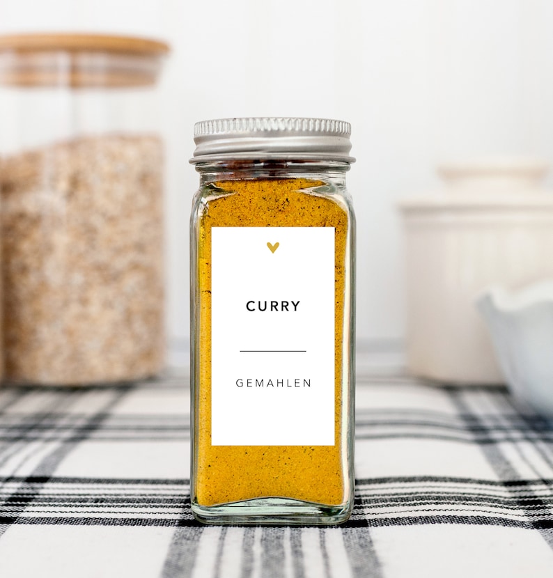 May include: A clear glass spice jar filled with yellow curry powder. The jar has a silver lid and a white label with the word "CURRY" and "GEMAHEN" printed in black. A small gold heart is above the word "CURRY". The jar sits on a black and white checkered surface.