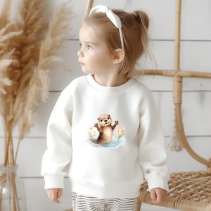 Könnte beinhalten: Ein weißes Sweatshirt mit einer Aquarellillustration eines Otters im Wasser mit Blumen. Der Otter ist braun und hellbraun, die Arme sind erhoben. Das Sweatshirt wird von einem kleinen Kind getragen.