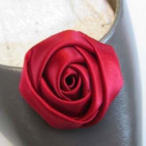 Pu&ograve; includere: Primo piano di un fiore di rosa in raso rosso. La rosa &egrave; su uno sfondo di tessuto grigio.