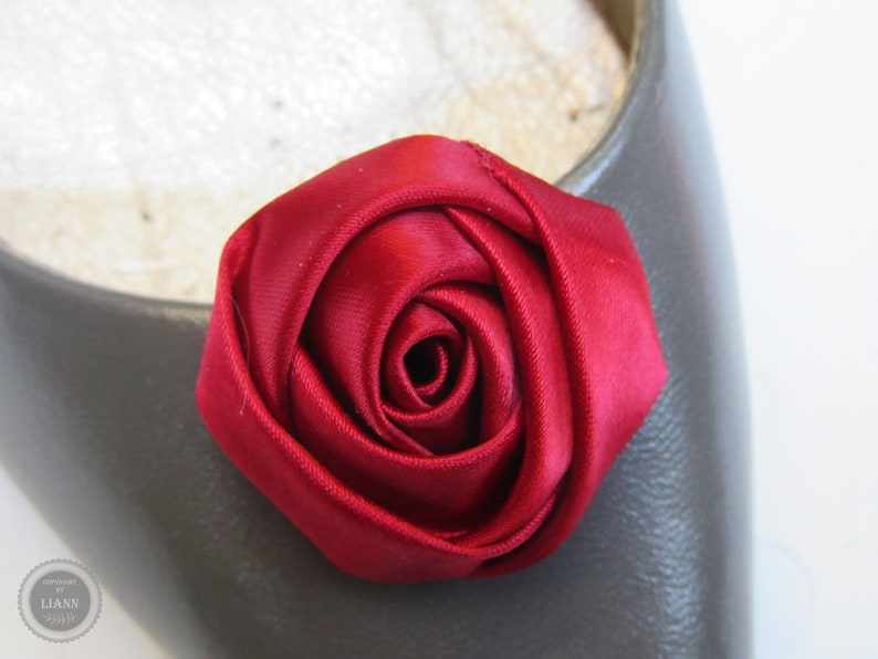 Pu&ograve; includere: Primo piano di un fiore di rosa in raso rosso. La rosa &egrave; su uno sfondo di tessuto grigio.