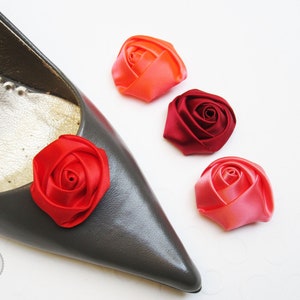 Pu&ograve; includere: Quattro decorazioni a forma di rosa in tessuto di raso in tonalit&agrave; di rosso, rosa e arancio. Le rose sono mostrate su una scarpa grigia e una superficie bianca.