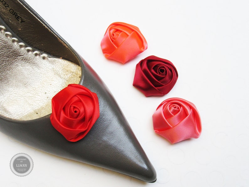 Pu&ograve; includere: Quattro decorazioni a forma di rosa in tessuto di raso in tonalit&agrave; di rosso, rosa e arancio. Le rose sono mostrate su una scarpa grigia e una superficie bianca.