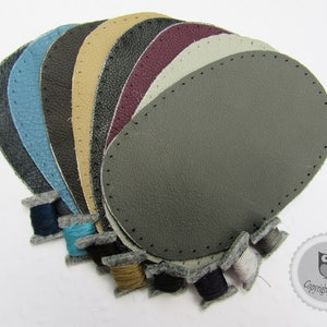 Könnte beinhalten: Eine Auswahl an ovalen Lederflicken in verschiedenen Farben, darunter Blau, Grau und Beige, jeweils mit einer genähten Kante. Kleine Garnrollen in passenden Farben sind darunter angeordnet. Die Flicken sind in einer fächerförmigen Anordnung.