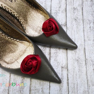 Pu&ograve; includere: Un paio di scarpe di raso grigio con decorazioni di rose in tessuto rosso sulle dita. Le scarpe sono su uno sfondo di legno bianco.