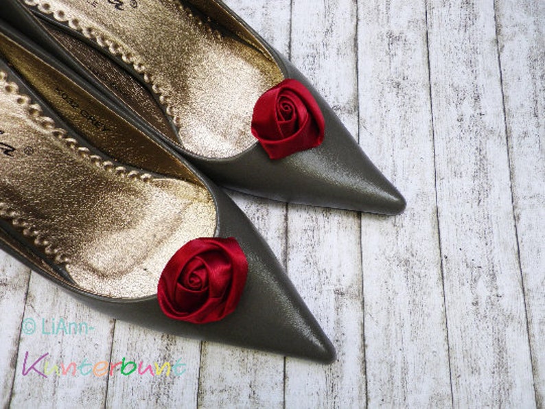 Pu&ograve; includere: Un paio di scarpe di raso grigio con decorazioni di rose in tessuto rosso sulle dita. Le scarpe sono su uno sfondo di legno bianco.