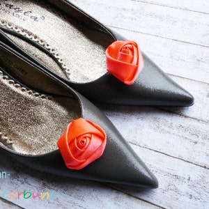 Pu&ograve; includere: Un paio di ballerine grigie con una rosa arancione decorativa su ogni scarpa. Le scarpe hanno un bordo dorato.