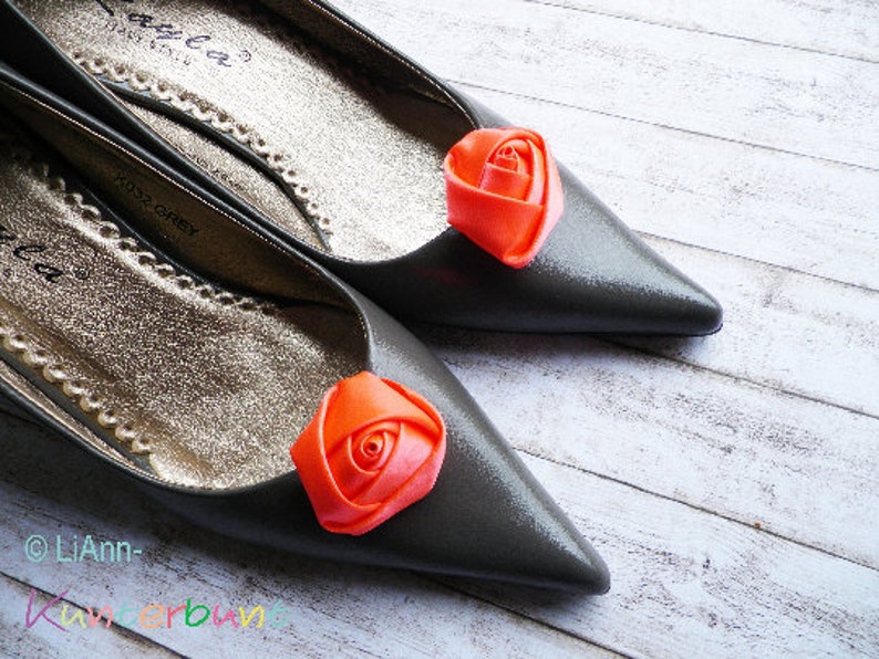 Pu&ograve; includere: Un paio di ballerine grigie con una rosa arancione decorativa su ogni scarpa. Le scarpe hanno un bordo dorato.