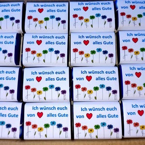 Könnte beinhalten: Eine Ansammlung von sechzehn rechteckigen Schokoladentafeln in weißem Papier. Jede Verpackung zeigt den blauen Text Ich wünsch euch von alles Gute, ein rotes Herz und eine Reihe bunter, illustrierter Blumen in Rot, Orange, Gelb, Grün, Blau und Violett.