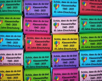Könnte beinhalten: Eine Ansammlung bunter Schokoriegel mit deutschem Text. Jeder Riegel trägt den Schriftzug "Schön, dass du da bist Klassentreffen 1945-2025 80 Jahre Einschulung" und eine Ballon-Grafik.