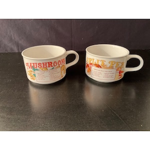 May include: Two white ceramic soup mugs with handles. One mug has "MUSHROOM" in red and the other has "SPLIT PEA" in yellow. Both mugs feature recipe text and illustrations in a vintage style.