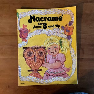 Könnte beinhalten: Ein Vintage-Macramé-Bastelbuch für Kinder ab 8 Jahren. Das Cover zeigt ein Cartoon-Mädchen in einem rosa Kleid, das eine Makramee-Eule hält. Das Buch hat einen gelben Hintergrund mit dekorativen Rändern und Illustrationen.