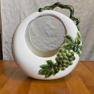 Può includere: Un cestino in ceramica bianca con un design circolare e un manico a forma di vite verde. Il cestino è decorato con uva e foglie verdi. Il cestino è su una superficie di legno.