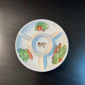 Puede incluir: Una fuente de cerámica blanca con tres secciones y un cuenco central para salsas. La fuente está decorada con ilustraciones coloridas de verduras, incluyendo zanahorias, alcachofas y champiñones. El borde tiene un borde azul claro.