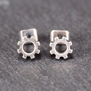 Può includere: Un paio di orecchini a lobo in argento a forma di ingranaggio. Gli orecchini sono piccoli e delicati e sarebbero un ottimo regalo per un meccanico o un ingegnere.