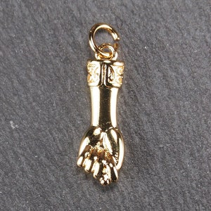 Könnte beinhalten: Goldfarbener Anhänger in Form einer Hand mit geschlossener Faust. Der Anhänger hat oben eine Schlaufe zum Anbringen an einer Halskette oder einem Armband.