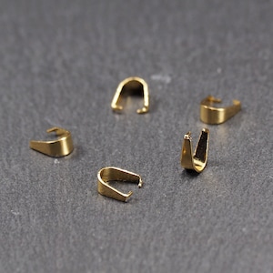 May include: Five gold-toned metal jump rings, also known as pendant bails, are arranged on a grey surface. These rings are used for attaching pendants to necklaces or bracelets.