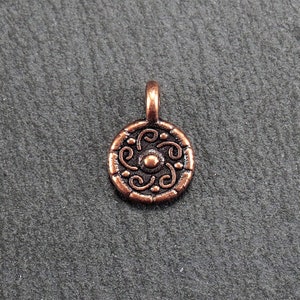 Op de afbeelding: Een koperkleurige hanger met een rond ontwerp met een centrale cirkel omgeven door ingewikkelde swirls en stippen.