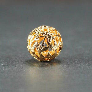 May include: A gold-colored metal bead with a delicate floral design. The bead is hollow and has a round shape.
