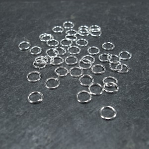 May include: A large quantity of silver jump rings, scattered on a dark grey surface. The rings are all the same size and are made of metal.
