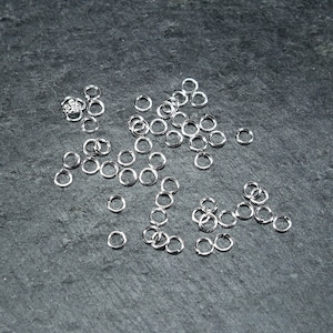 May include: A large quantity of silver jump rings scattered on a dark grey surface. The jump rings are small and circular, perfect for jewellery making.
