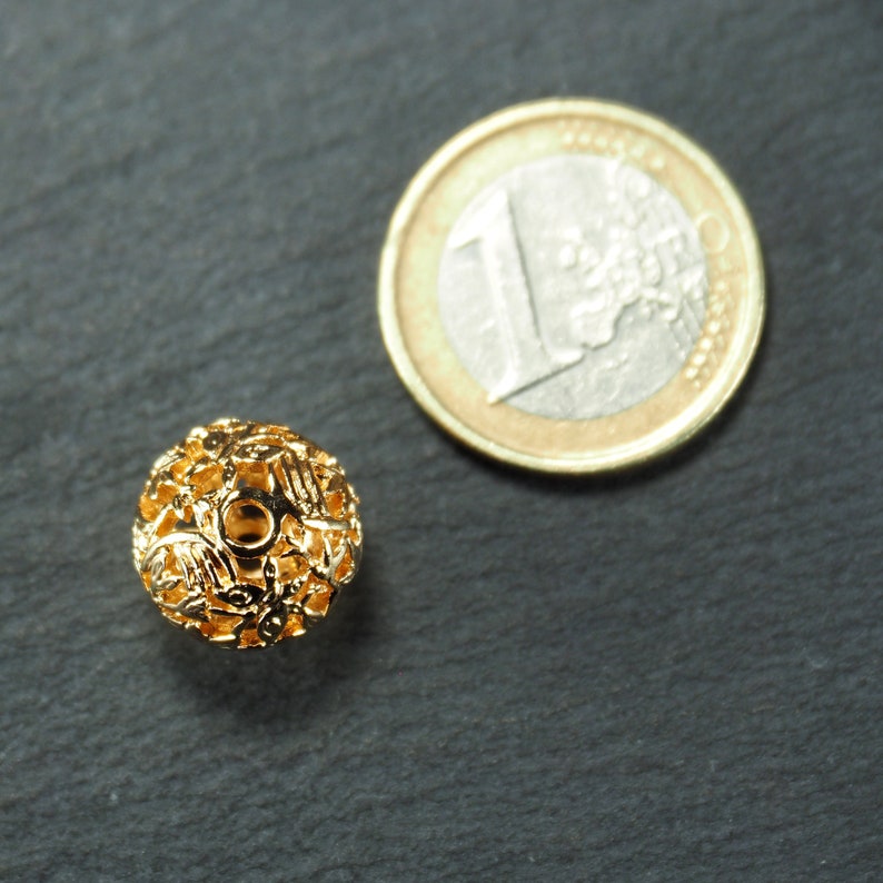 May include: A gold-colored metal bead with a decorative, openwork design. The bead is about the size of a US one-euro coin.